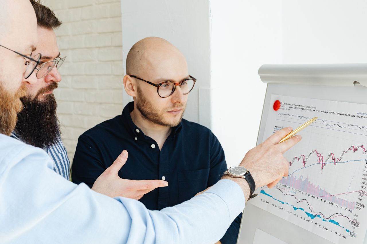Three men discussing financial charts on a whiteboard during a business meeting.